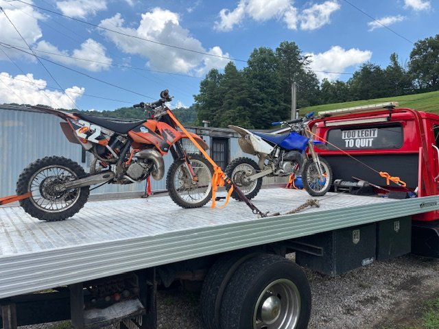 Two dirt bikes, orange and blue, secured on a red tow truck bed under a cloudy sky.