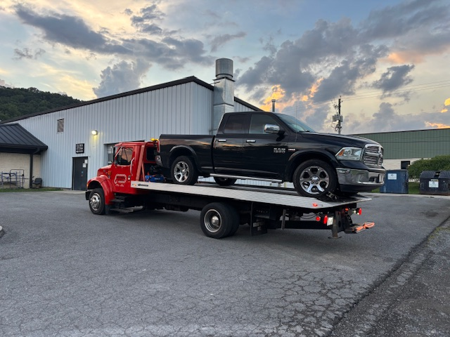 Red tow truck carrying a black pickup truck on a flatbed in a parking lot.