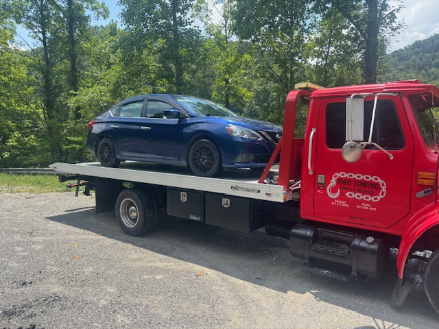 Blue sedan on a red tow truck bed, parked on gravel with trees in the background.
