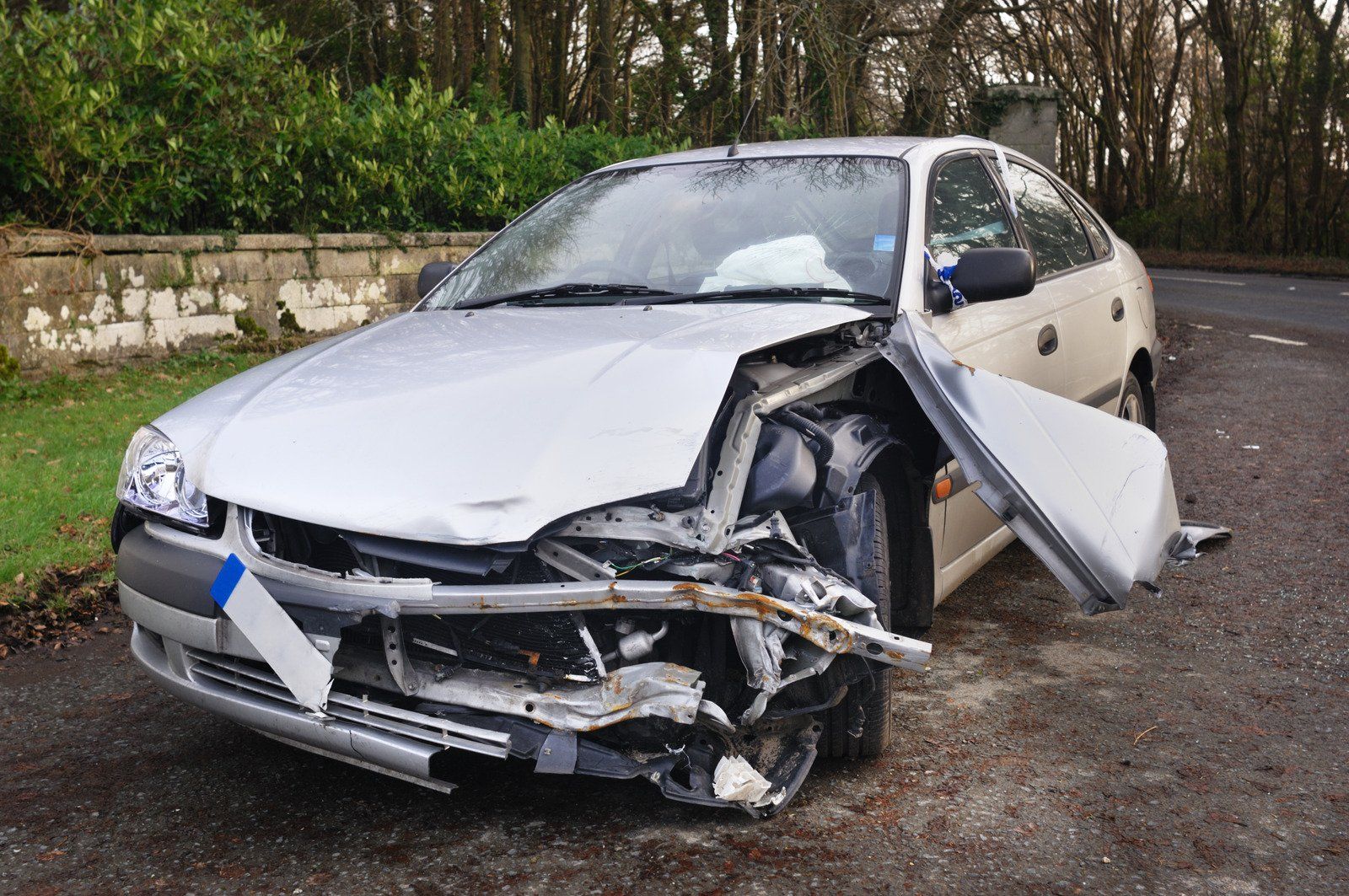 Damaged silver car after a collision, front end crumpled, parked on the side of a road.