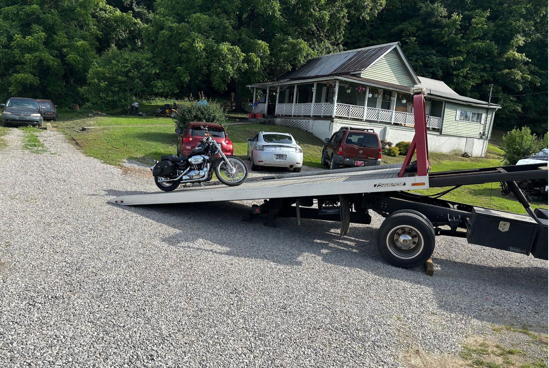 Motorcycle on a flatbed truck, parked in front of a house on a gravel driveway.
