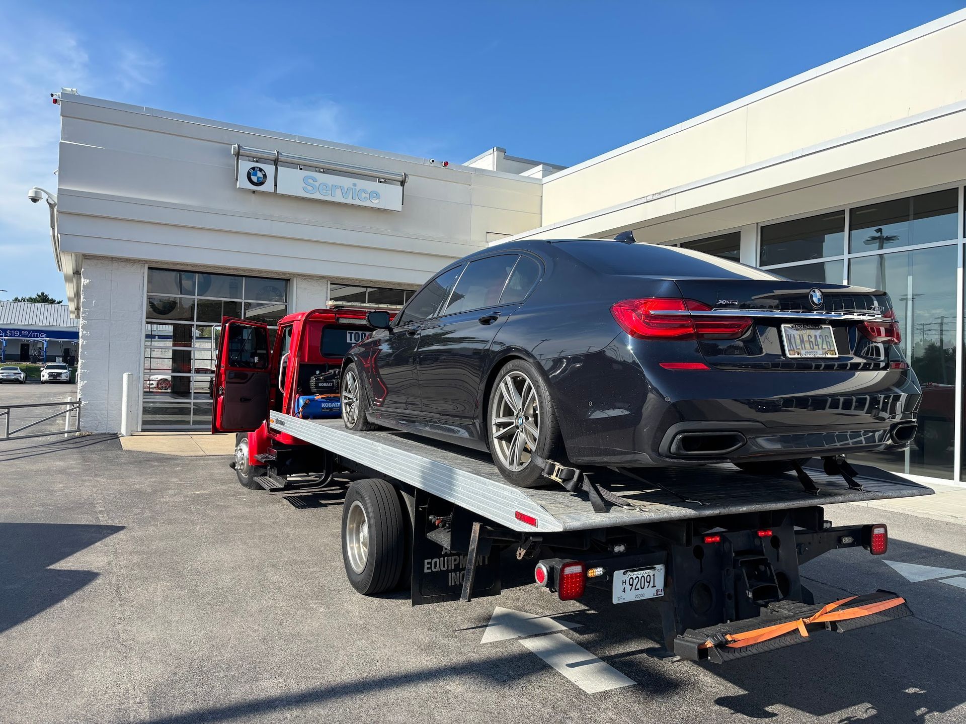 A dark BMW sedan being towed on a flatbed tow truck in front of a car dealership.
