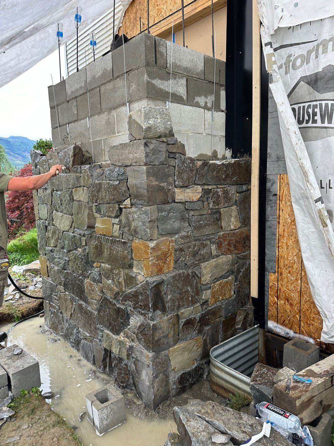 Construction of a stone and concrete block corner wall on a residential building exterior. Person's hand visible.