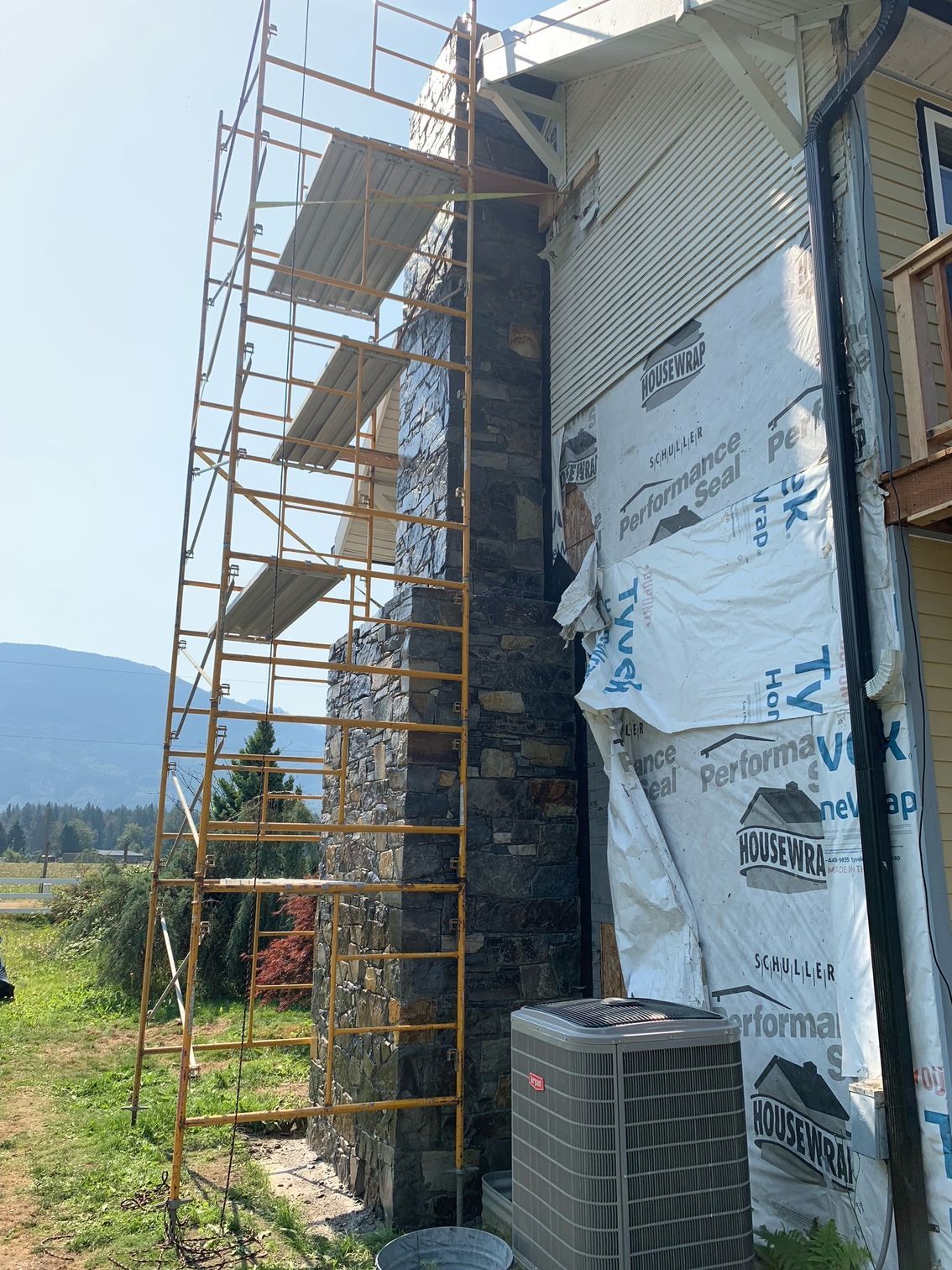 Scaffolding next to a stone chimney being worked on, set against a blue sky and mountain view.