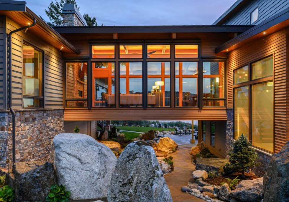 Modern home with a walkway over a rock garden; large windows, warm interior lighting.