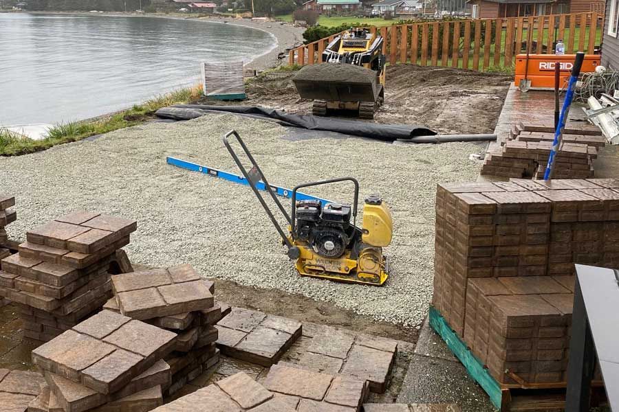 Construction site: paving stones being laid near a beach; a compactor and mini-excavator are used.