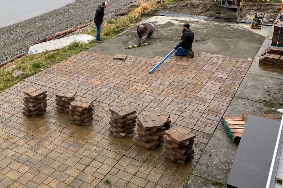 Workers installing brown patio tiles near a body of water. Piles of tiles sit nearby.