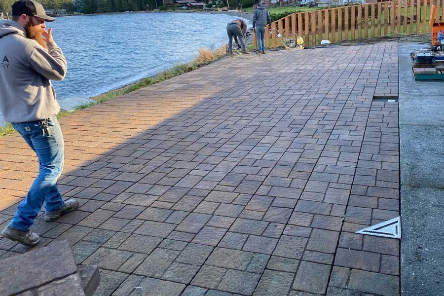 Man looking distressed on a brick patio next to a lake; two other men work in background.