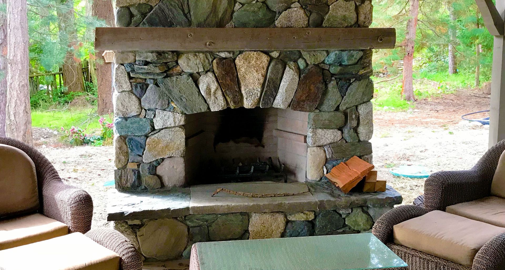 Stone fireplace on a patio with wicker furniture and a wooded background.