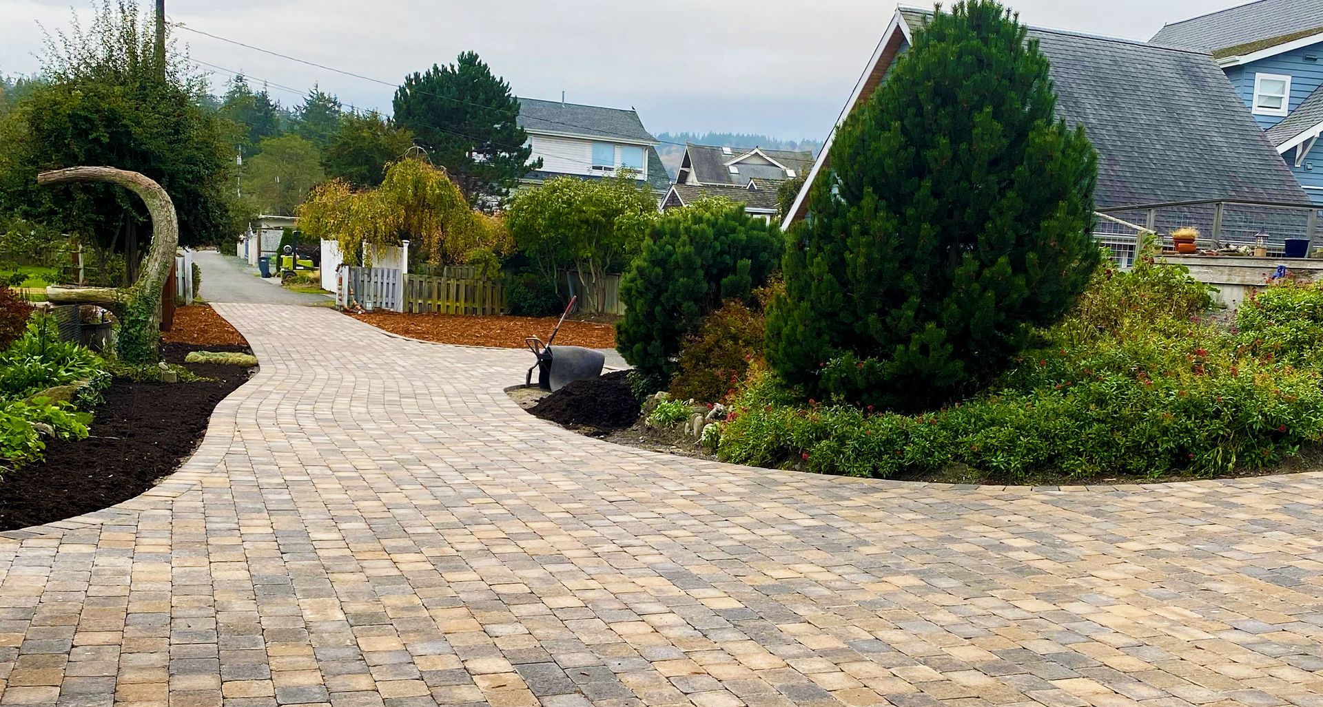 Brick paved driveway leading toward houses, surrounded by greenery and a white picket fence.