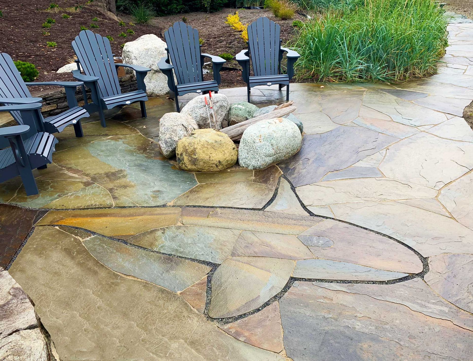 Patio with stone inlay of an orca. Gray Adirondack chairs, rocks, and green foliage.