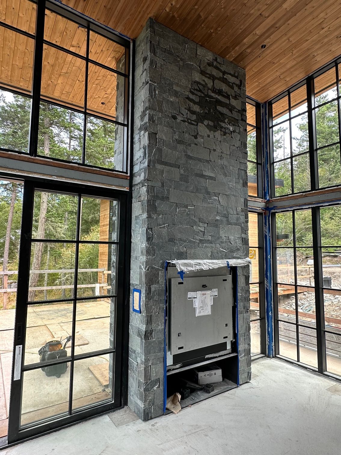 A tall, grey stone fireplace in a room with large windows, concrete floor, and wood ceiling.