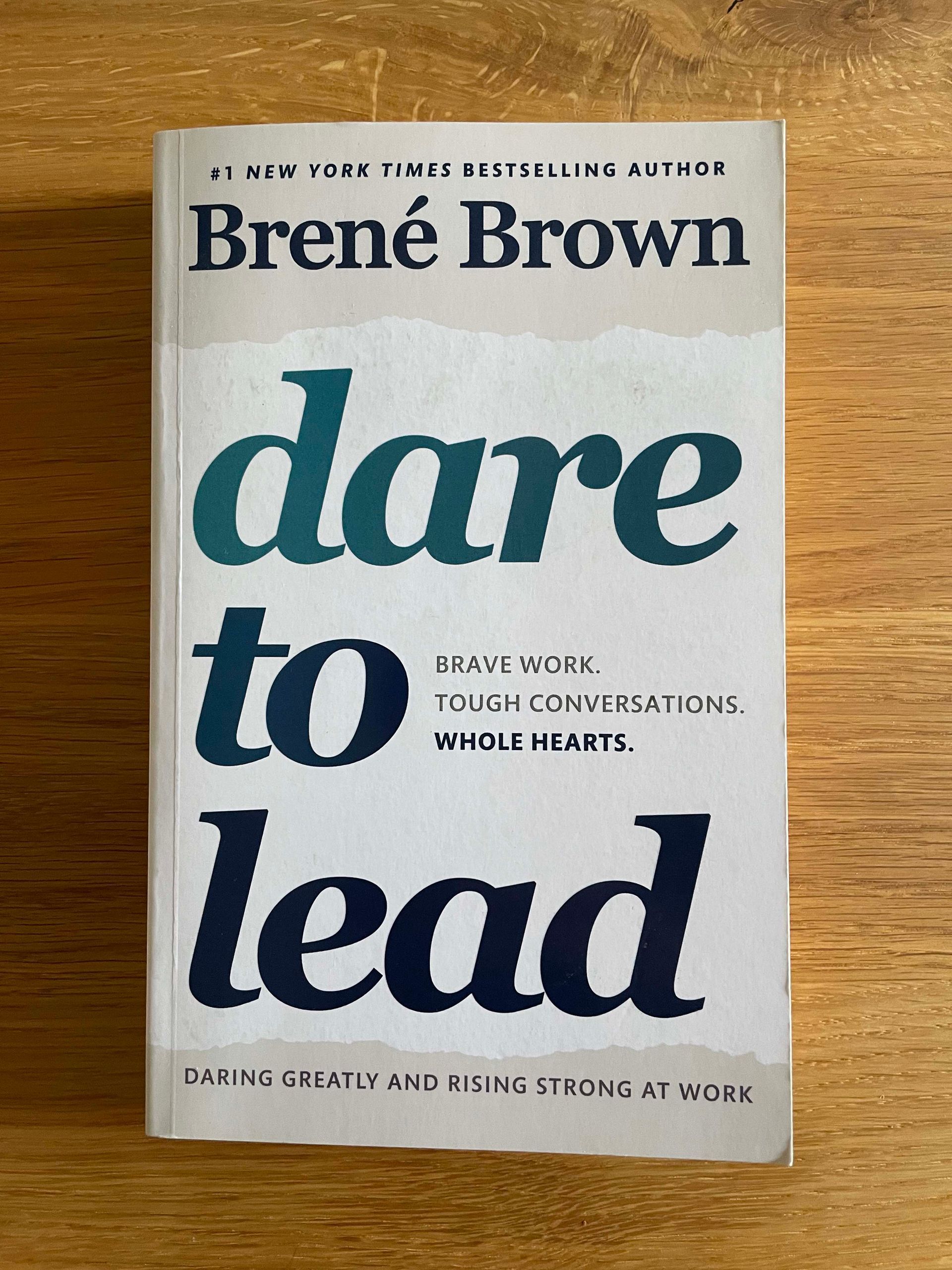 Picture of the book cover of dare to lead by Brené Brown on a wooden surface. The book cover is white and grey with a dark blue font with gradient. The subtitle is: brave work, tough conversations, whole hearts.