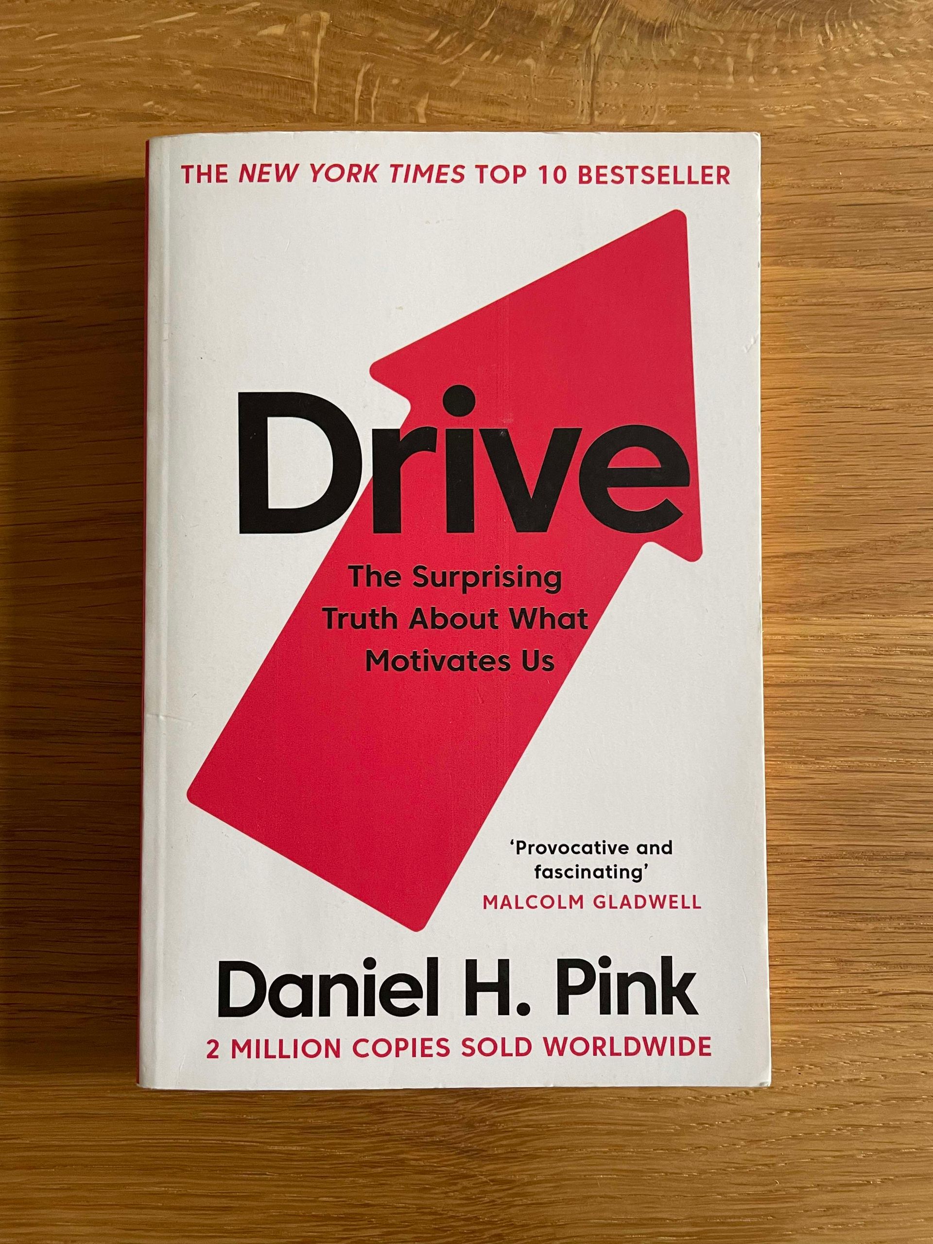 Picture of the book cover of drive by Daniel H. Pink on a wooden surface. The book cover is white with a red arrow pointing upwards. The font is black and red. The subtitle is: the surprising truth about what motivates us.