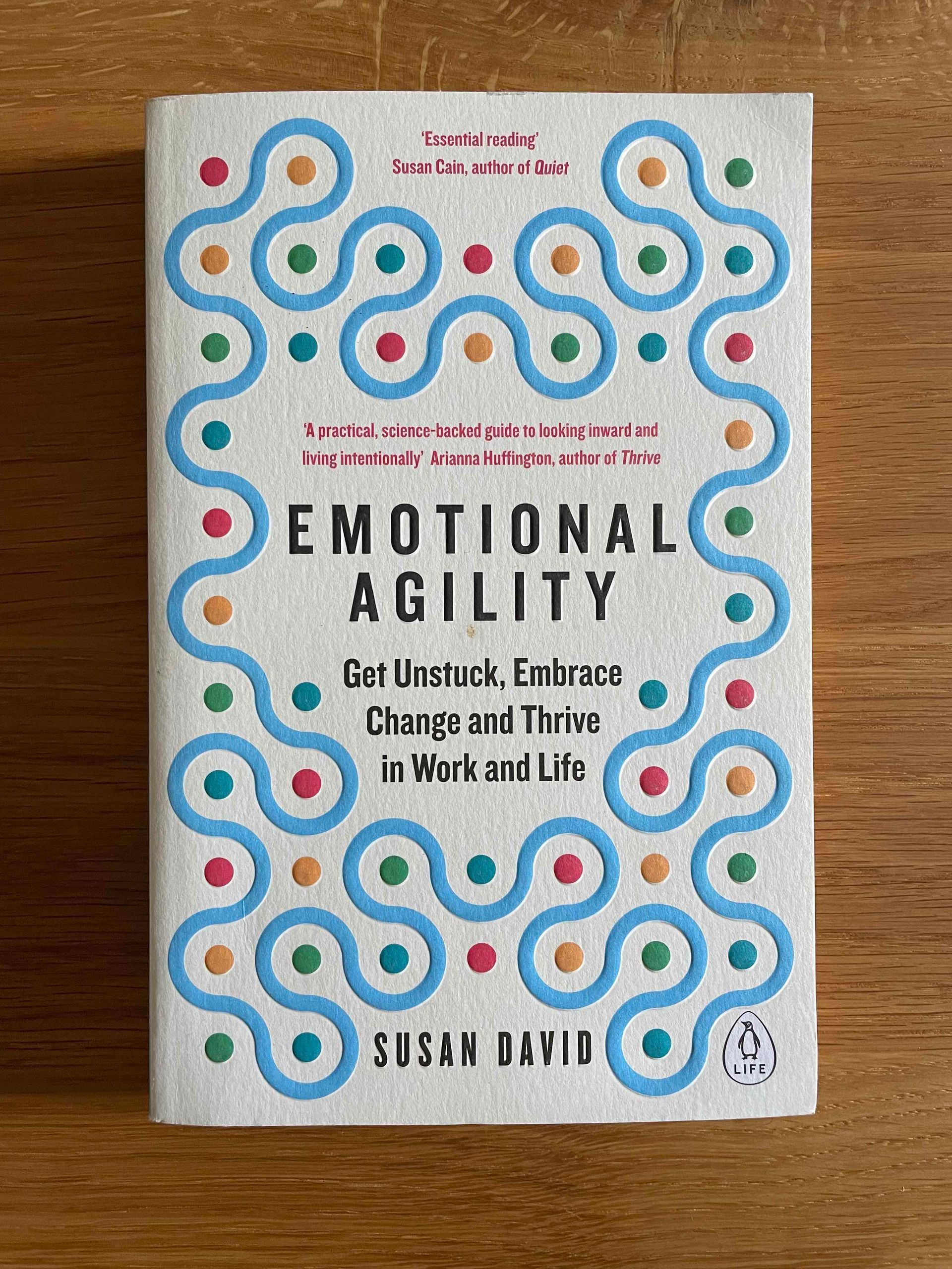 Foto des englischen Buchcovers von Emotional Agility von Susan David auf einer Holzoberfläche. Das Buchcover ist weiß mit einer blauen, welligen Linie und roten, grünen, blauen und gelben Punkten. Die Schrift ist schwarz und rot. Der Untertitel lautet: get unstuck, embrace change and thrive in work and life.