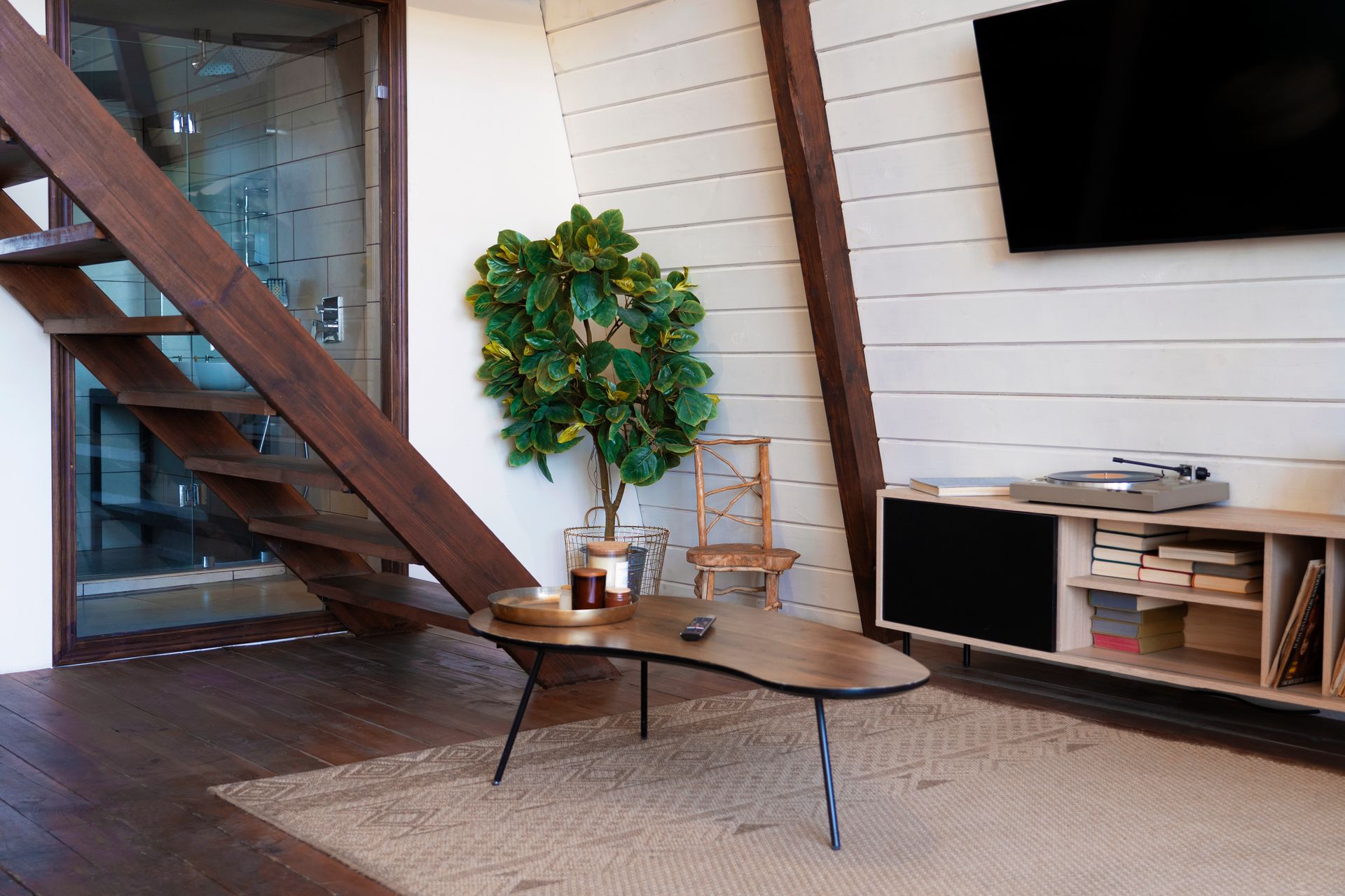 A modern living room with a wooden staircase, a kidney-shaped coffee table, a potted plant, and a TV on a console table.