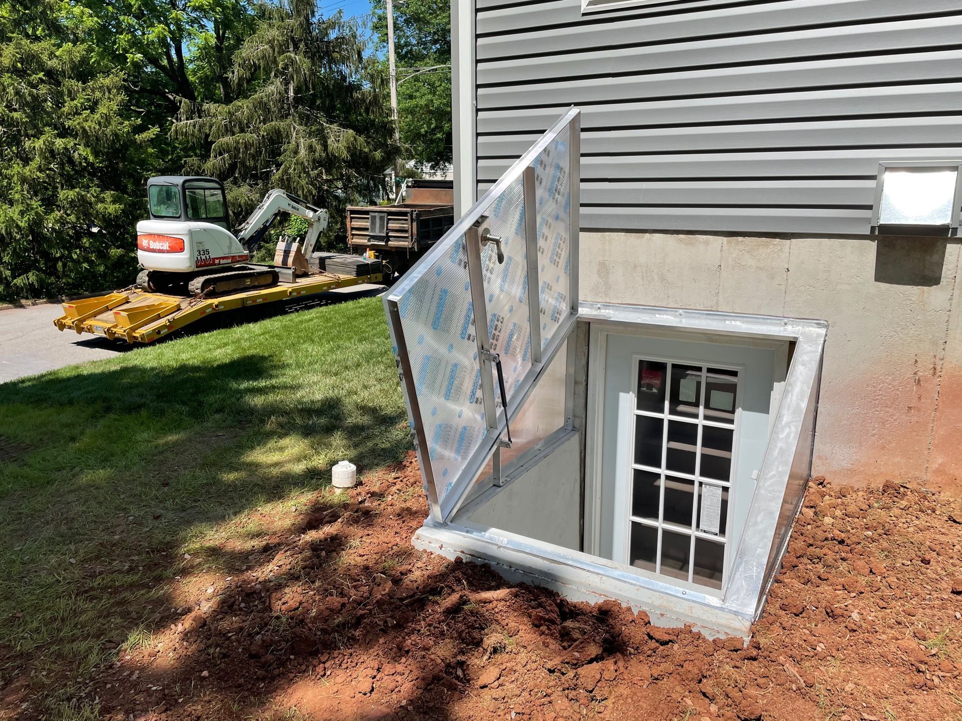 An open metal bulkhead cellar door leads to a white door in the foundation of a house next to a parked construction trailer.