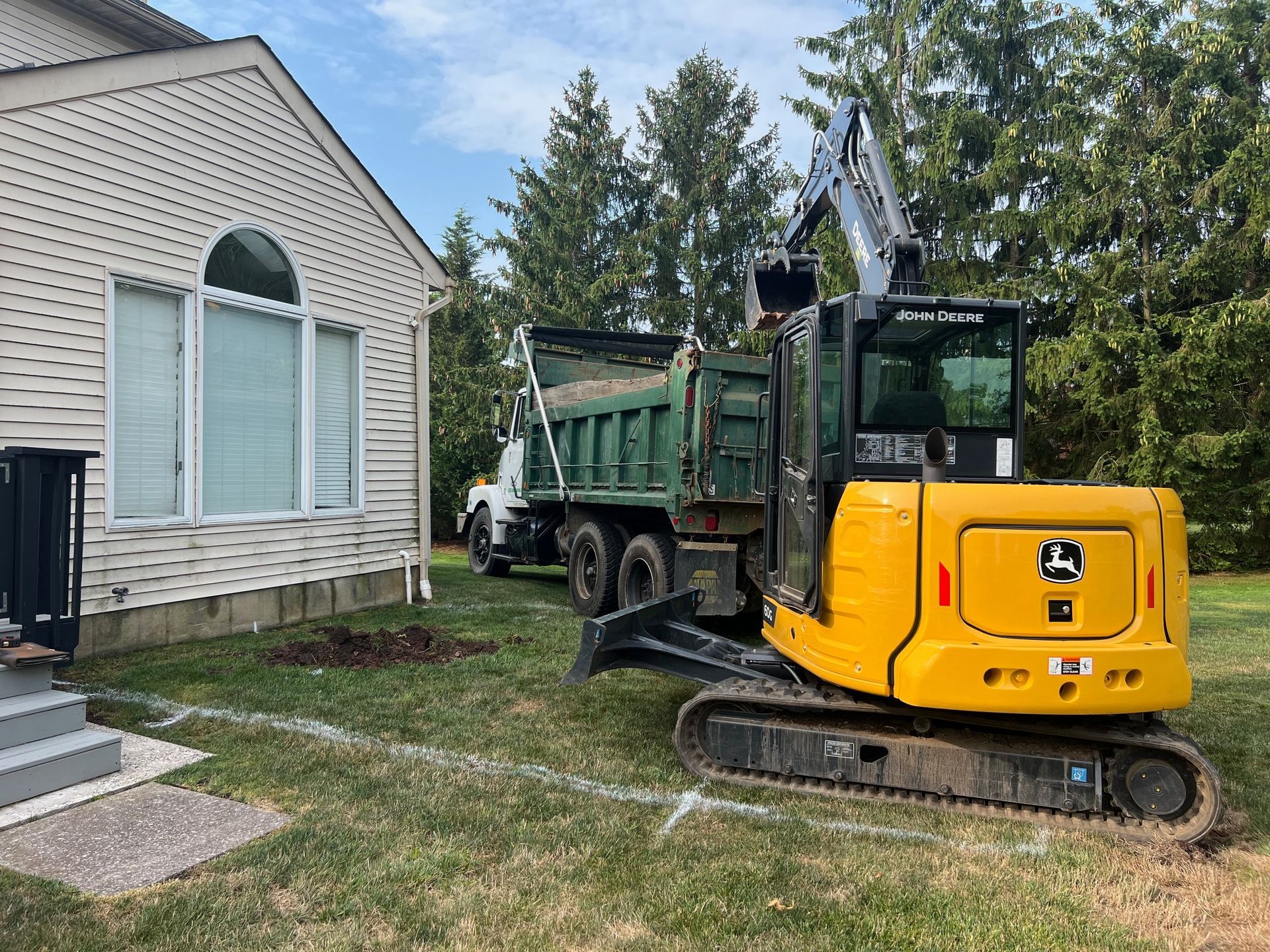 Yellow excavator loading a green dump truck next to a house on a lawn.