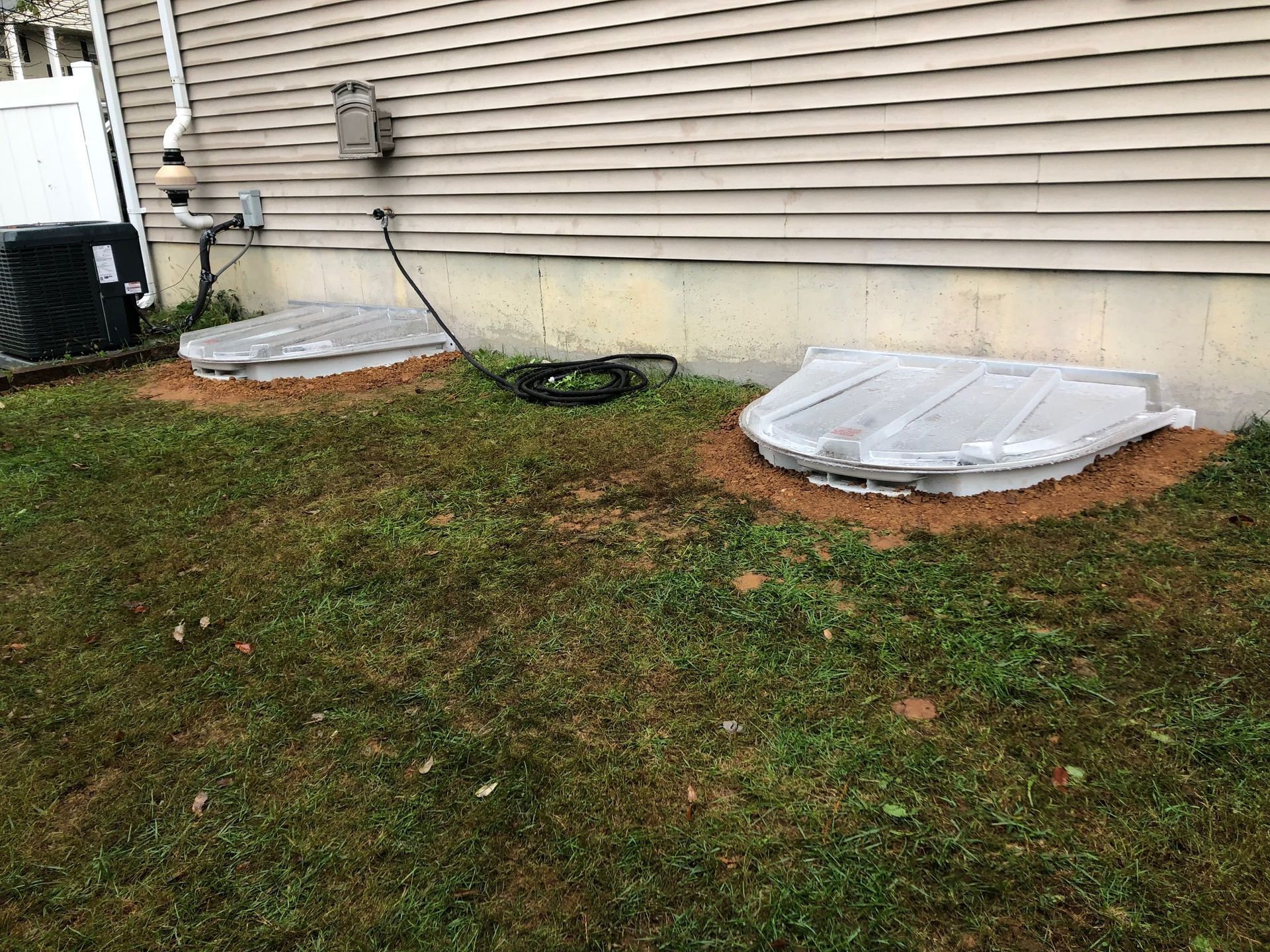 Two white plastic window well covers sit on the ground against a beige vinyl-sided house foundation.