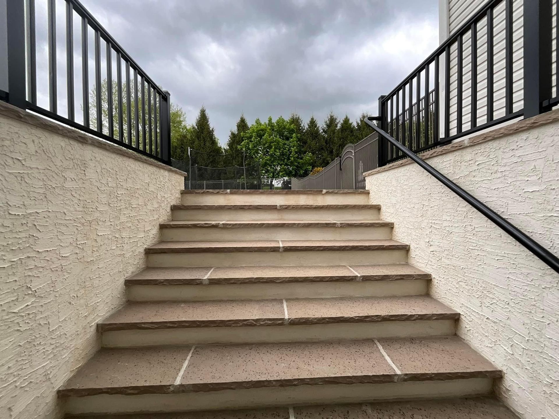 A set of outdoor concrete stairs with black railings on both sides, leading up toward trees against a cloudy sky.