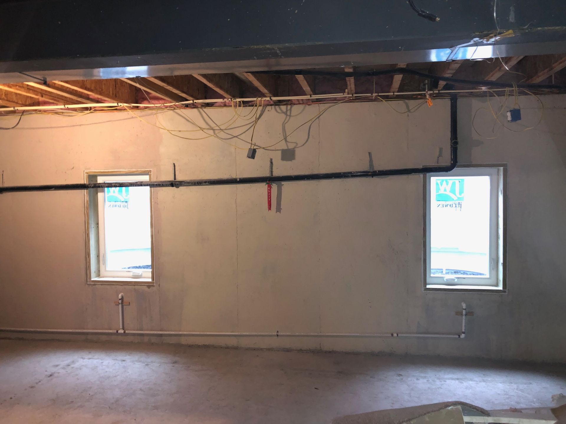 Unfinished basement wall with two windows, exposed ceiling joists, and various pipes and wires running across it.