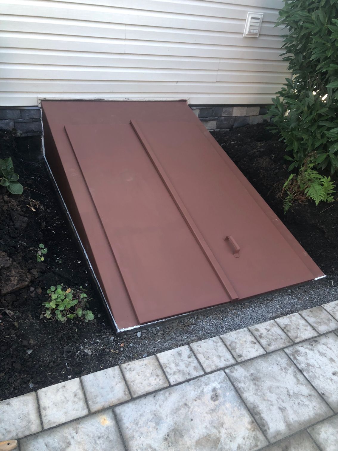 Brown basement door set into the ground against a siding wall.