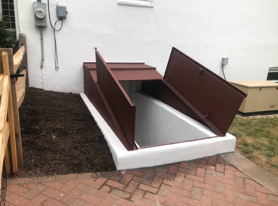 Brown metal bulkhead basement doors open on a white concrete foundation against a white wall next to a brick patio.
