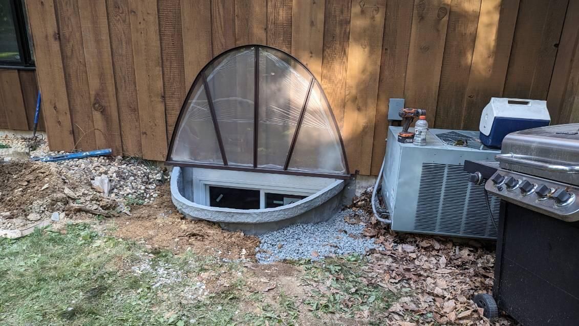 A basement window well with a clear, arched plastic cover installed next to a wood-sided house and an AC unit.