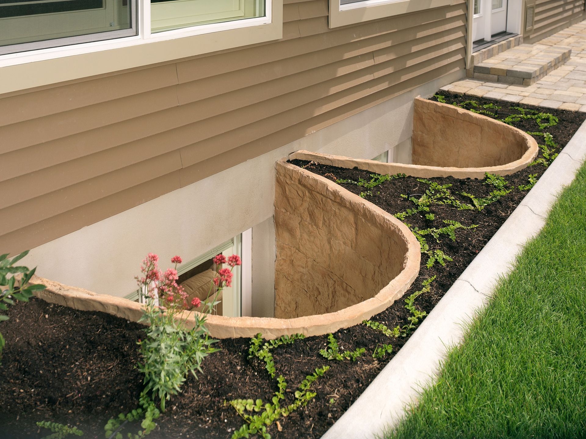 Curved window wells with landscaping against a tan house, surrounded by grass and a walkway.