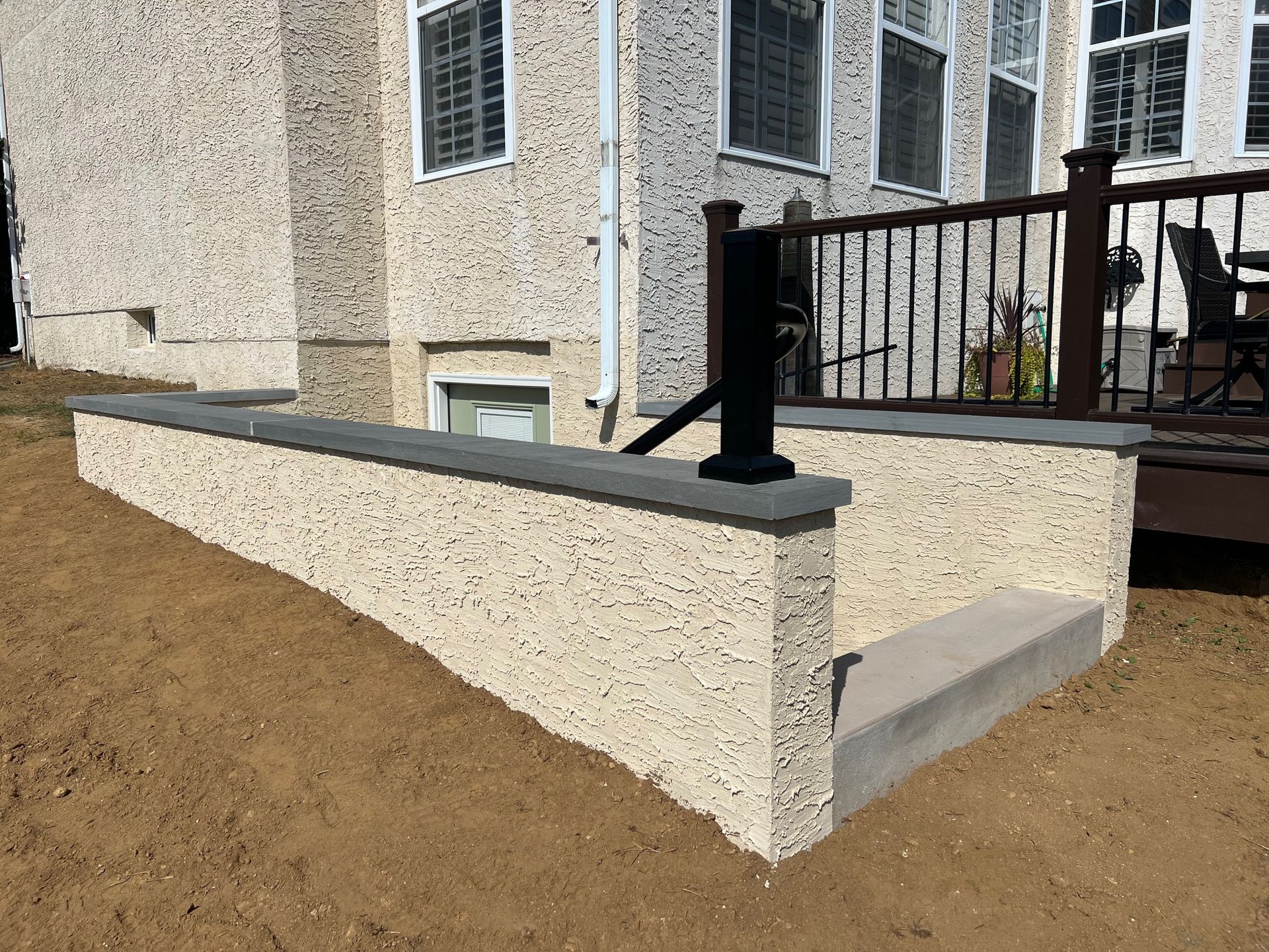 A beige masonry wall bordering outdoor concrete stairs next to a house with a dark railing and a deck above.