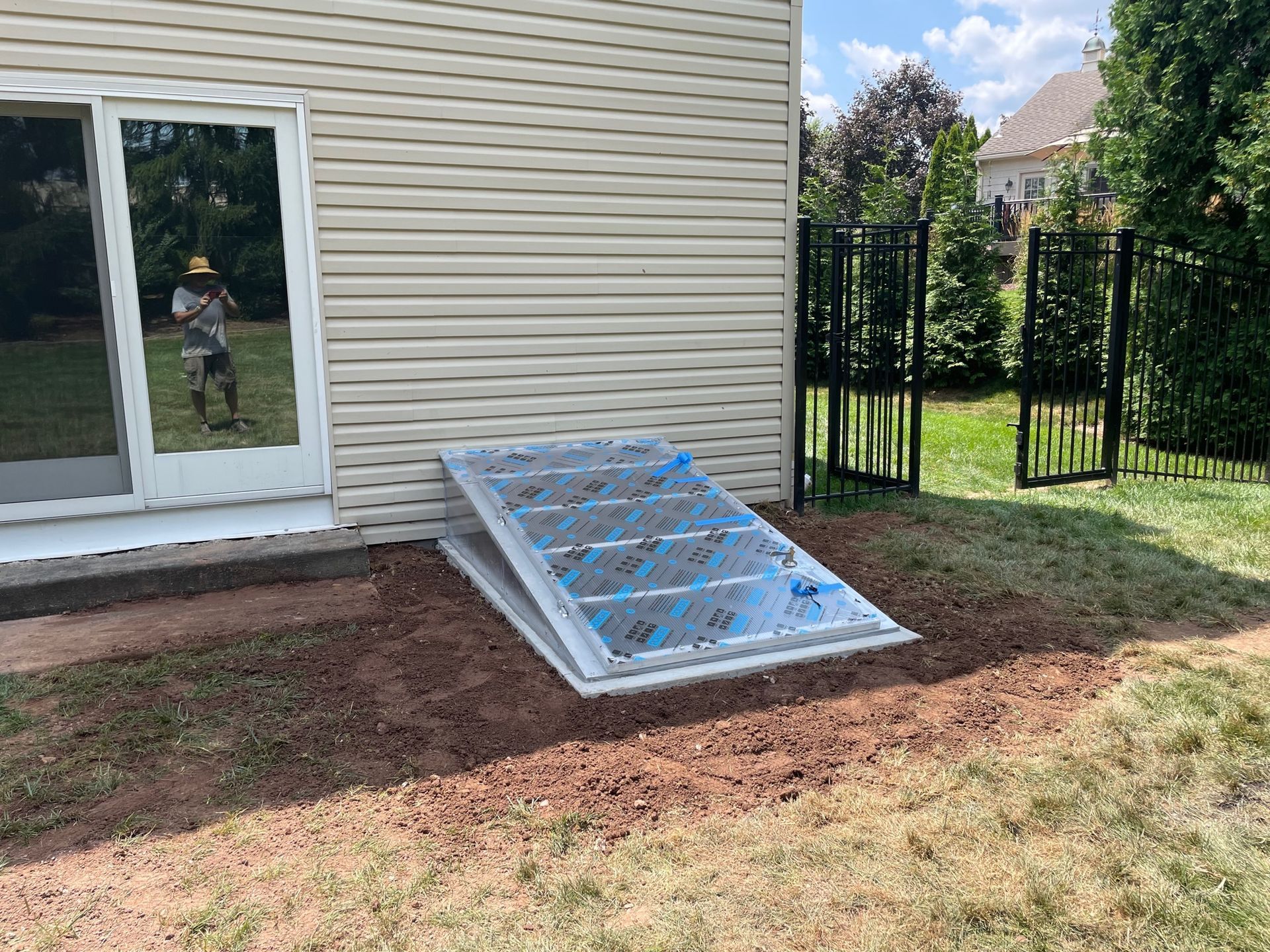 A basement window with a transparent cover next to a sliding glass door and a black fence on a grassy lawn.