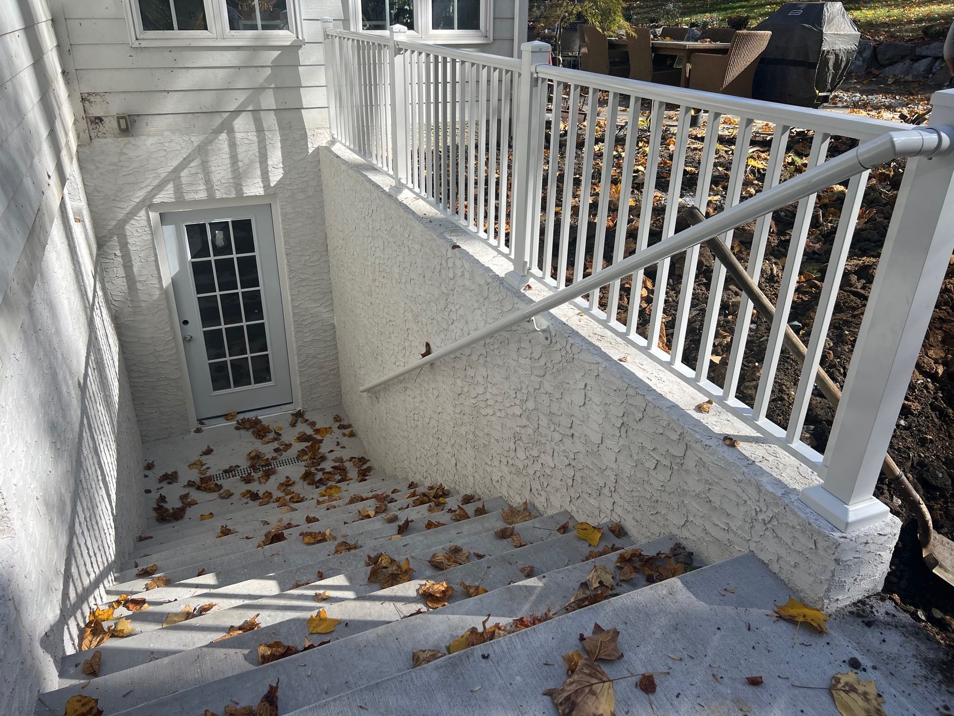 White concrete outdoor stairs descending to a basement door, featuring a white metal railing and scattered autumn leaves.