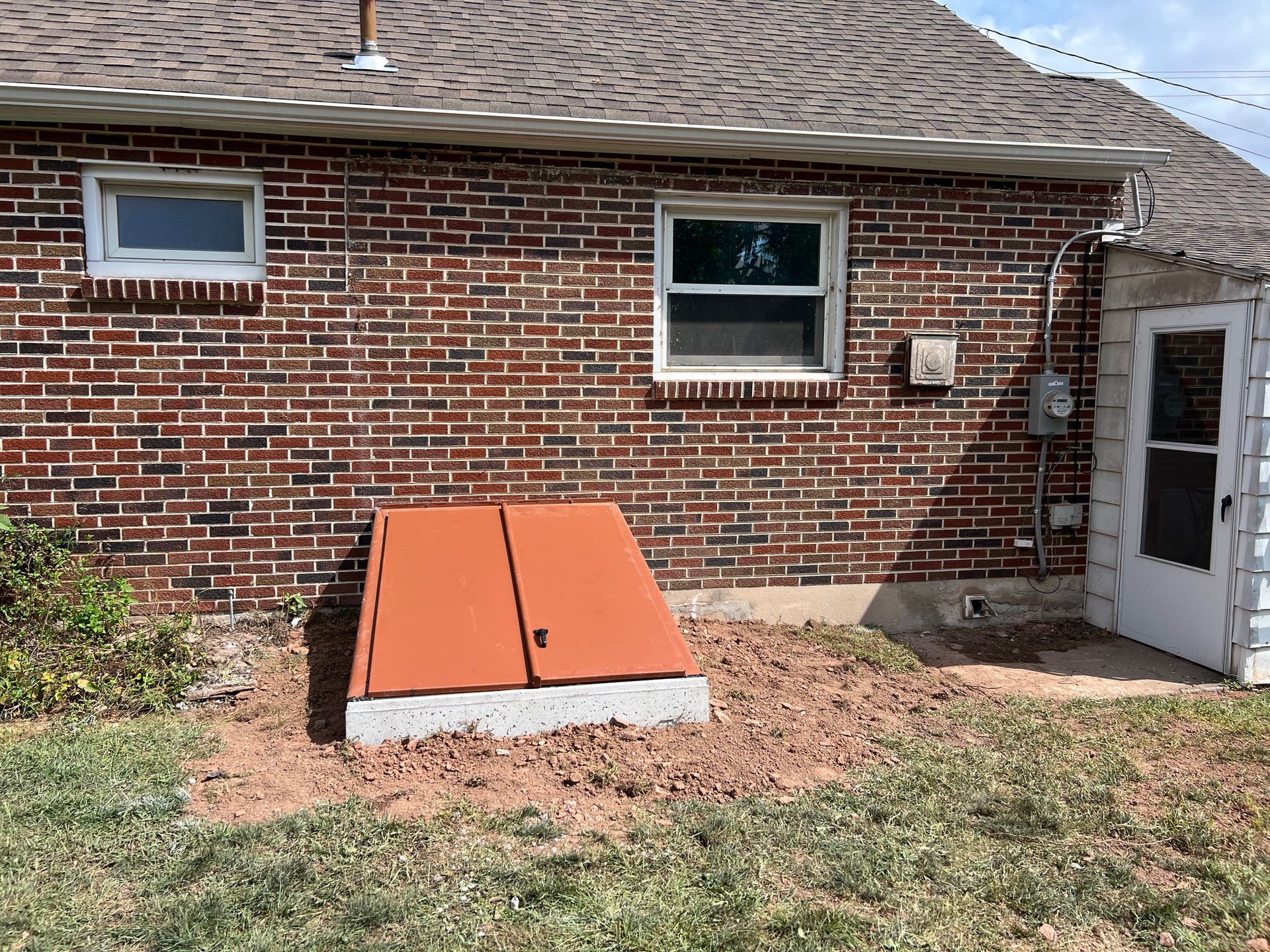 Exterior of a brick house featuring a newly installed orange metal bulkhead basement door over a concrete foundation.