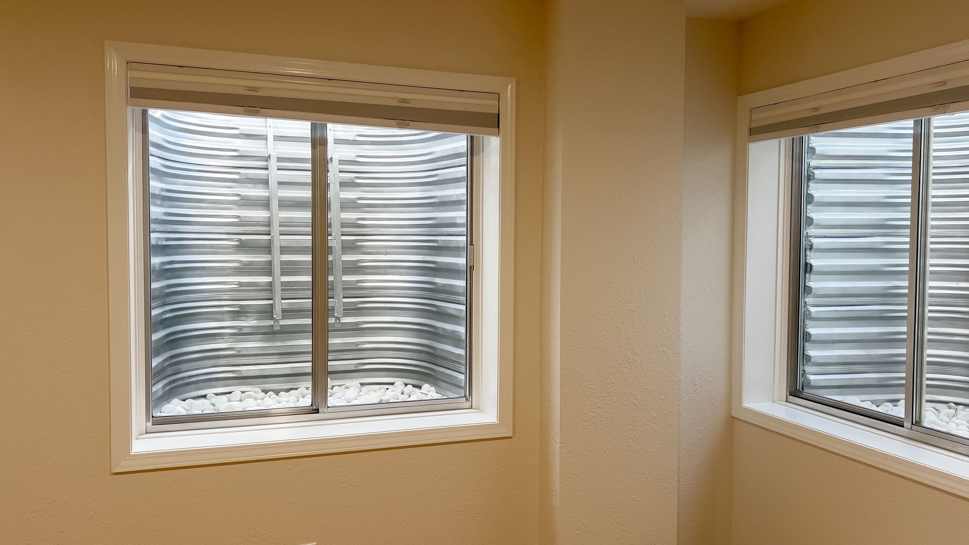 Two basement windows with corrugated metal wells, inside a light-colored room.
