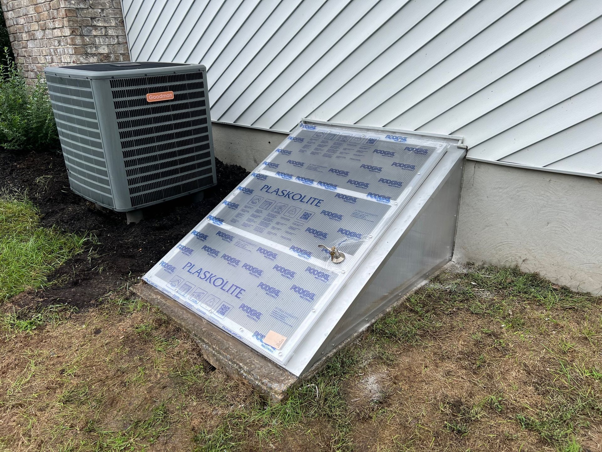 A metal window well cover next to an AC unit and a house, in a grassy yard.