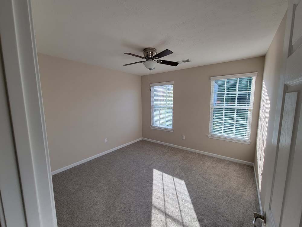 An empty bedroom with two windows and a ceiling fan.