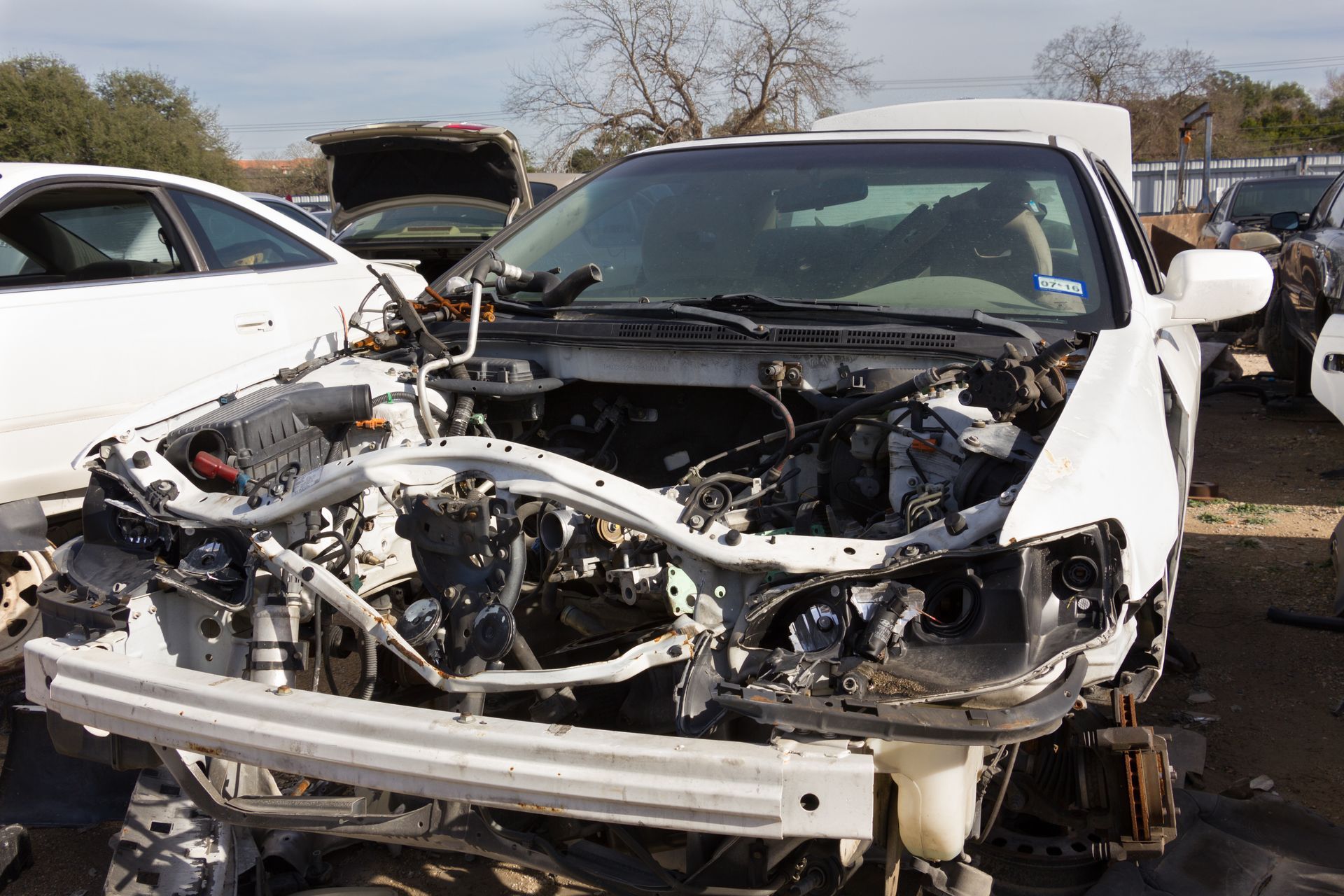 A white car with the hood open is sitting in a junkyard.
