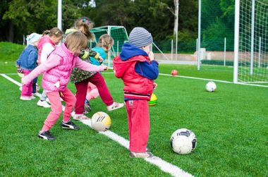 Children in colorful sportswear practice soccer drills on a green field near the goal.