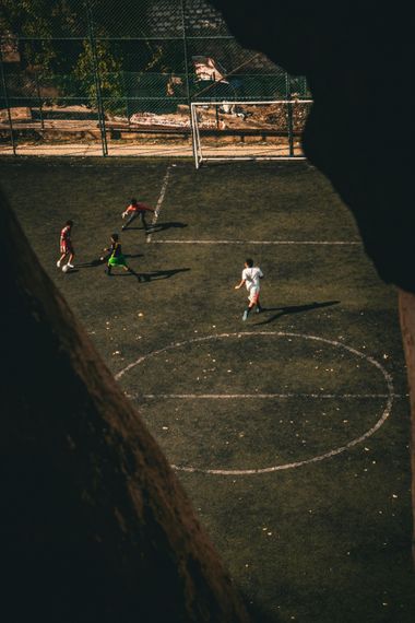 Four players on a small outdoor soccer court, with a goal at the top in dim light.