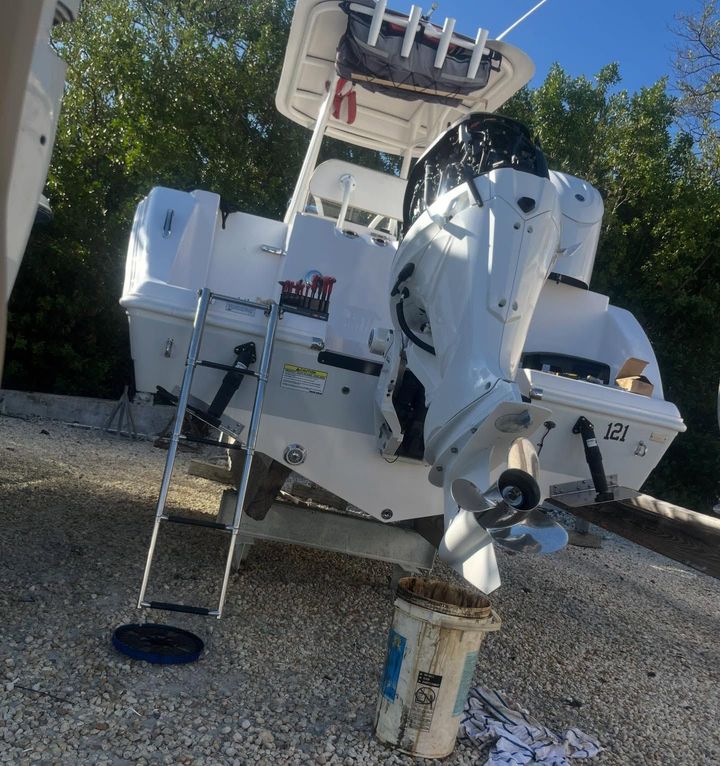White boat with two outboard motors on a gravel surface. A ladder is leaning against the side.