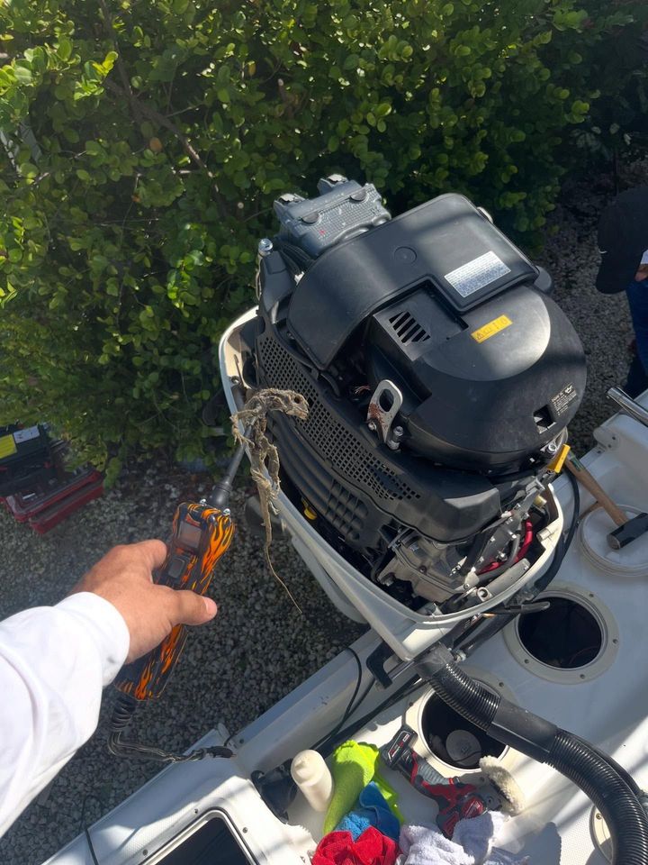 Man working on a white outboard motor attached to a boat. The motor is black and gray. Tools and vegetation are visible.