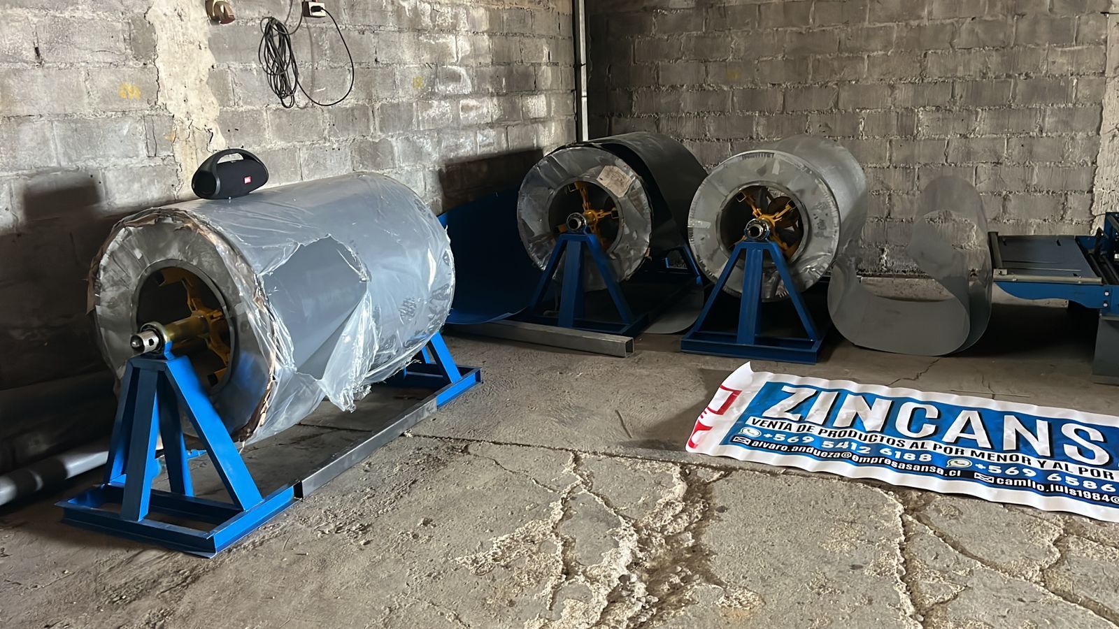 Rollos de láminas de metal gris sobre soportes azules en una habitación con paredes de ladrillo con un cartel que dice