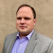 A man in a suit and purple shirt is standing in front of a wall.