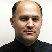 A bald man wearing a black shirt and tie is looking at the camera.
