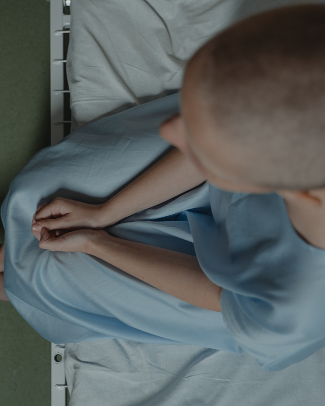 Bald person in a blue gown sits on a hospital bed, hands clasped, looking down.