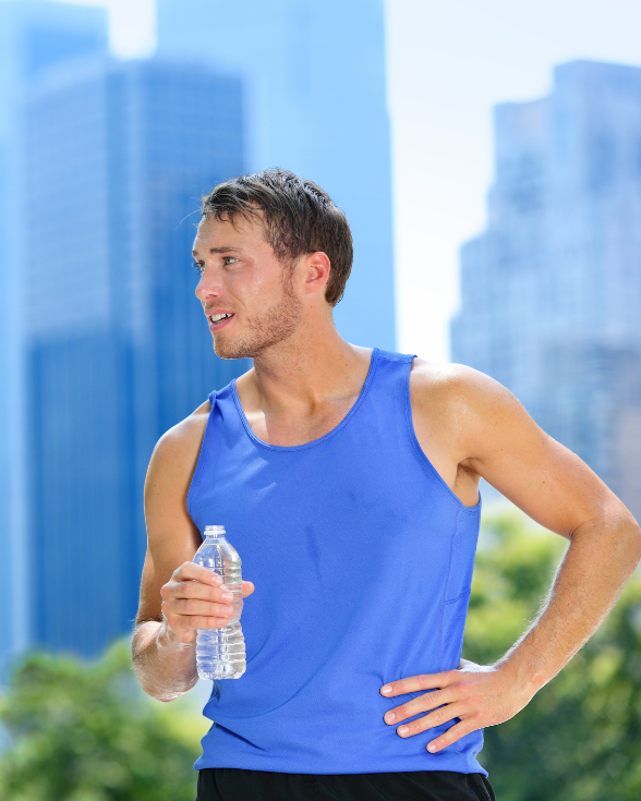 Man in blue tank top holding water bottle, looking to the side, in front of city buildings.