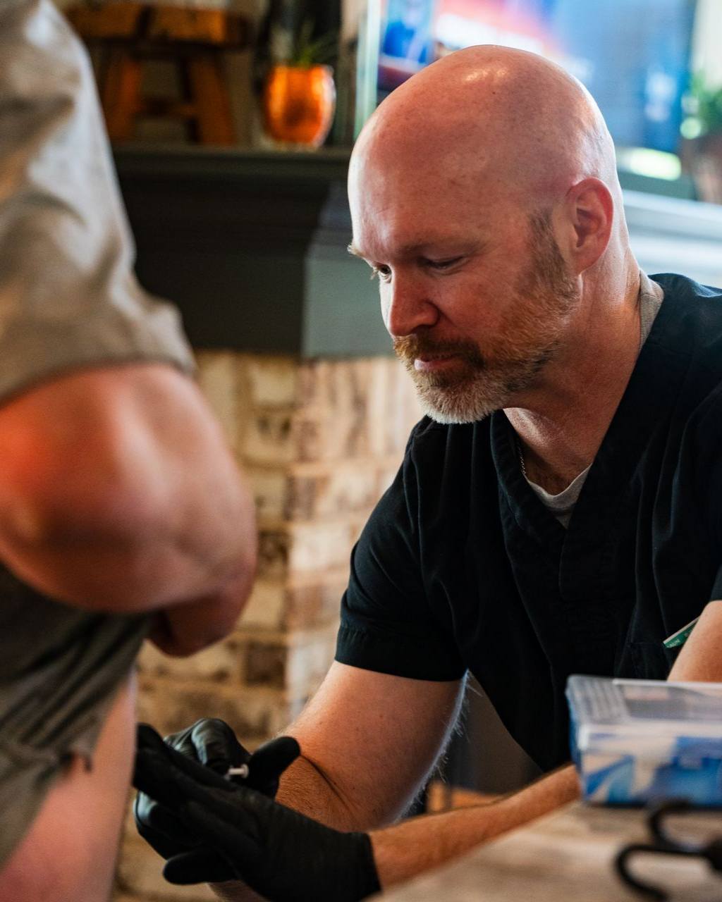 Man wearing black gloves focused, examining something held near another person's body. Brick wall in background.