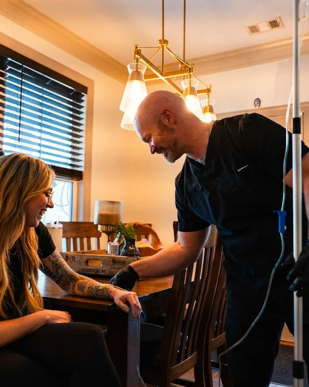 A person giving an IV drip to a smiling person seated at a table. Indoors, warm lighting.