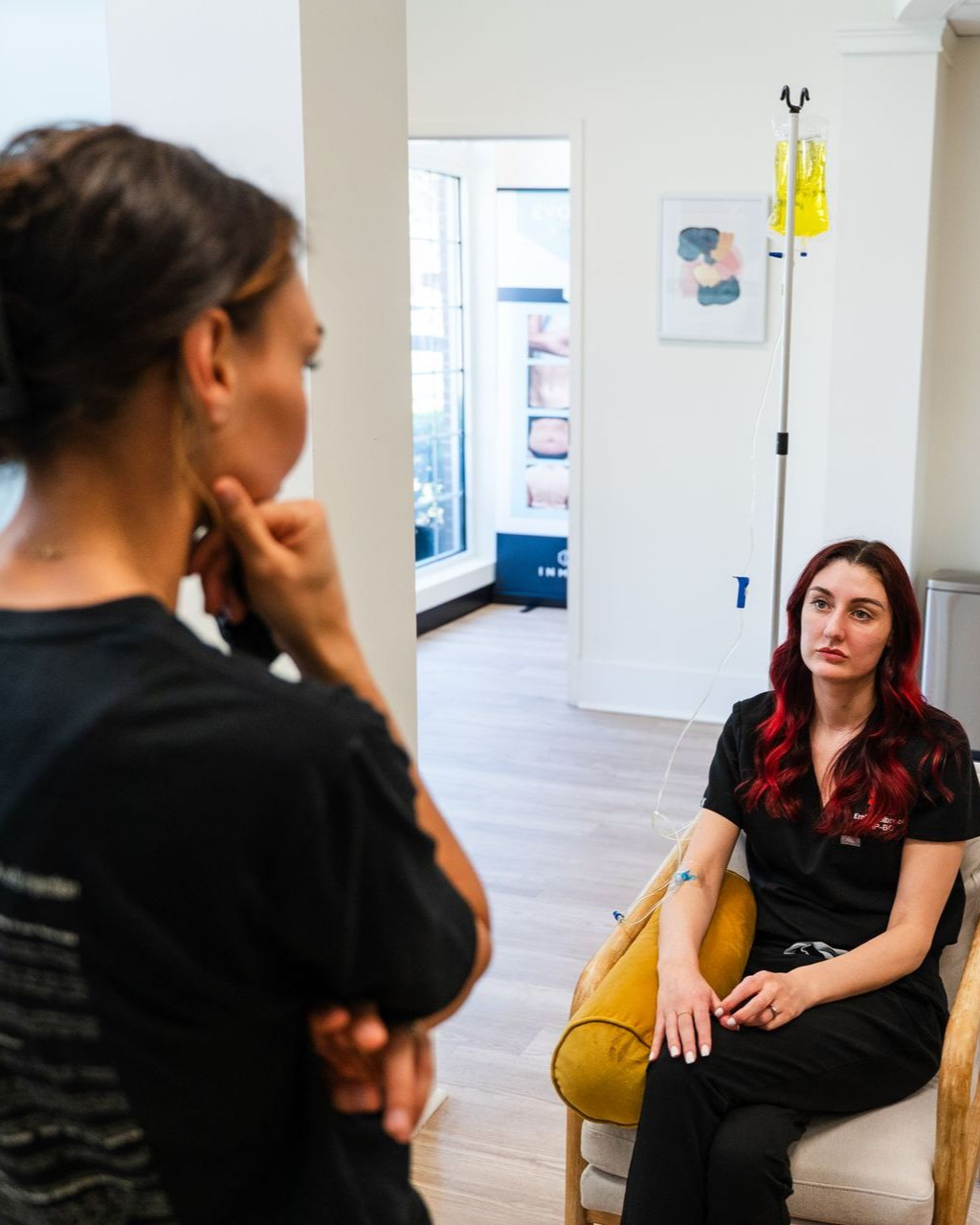 Woman talks to seated person with IV drip in a modern medical setting.