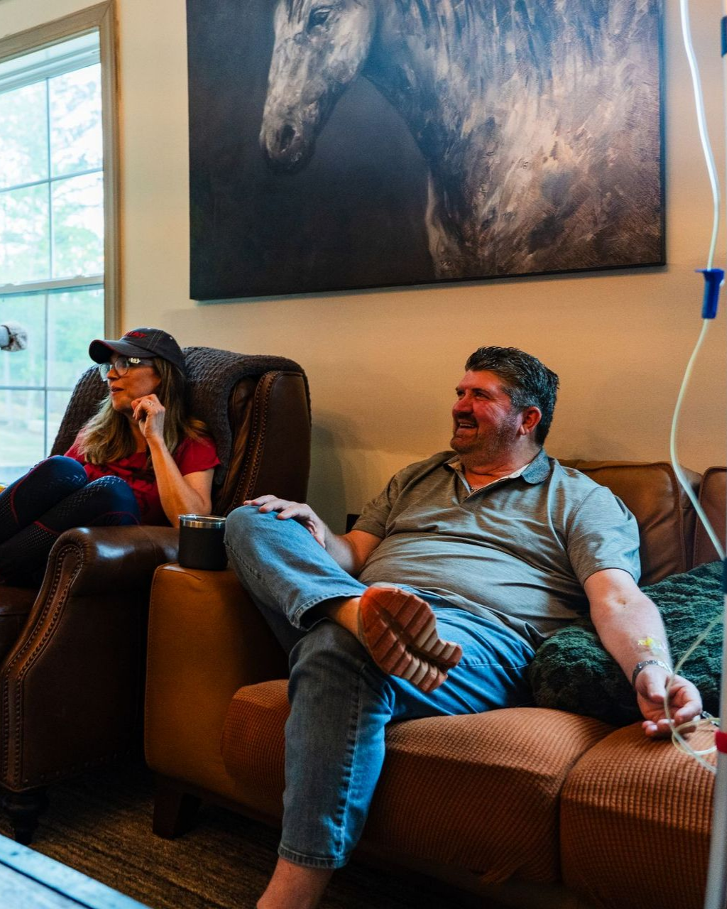 Two people relax on leather sofas, one with an IV drip, beneath a horse portrait in a living room.